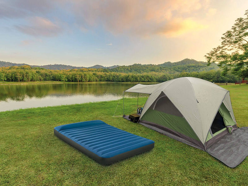 Laad afbeelding in galerijtoner, Een blauw Intex TruAire opblaasbaar twijfelaar kampeerluchtbed naast een opgezette tent beide staan op het gras aan een meer