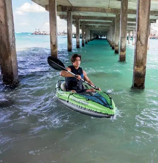 Een persoon kajakt onder een betonnen pier in de Intex Challenger K1 Opblaasbare Kajak 1-persoons van Intex, glijdend door helder turquoise water - ideaal voor avontuurlijke tochten op zonnige dagen.