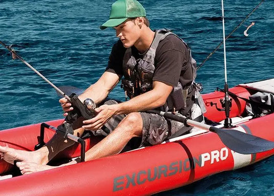 Man in een Intex Excursion Pro K1 Opblaasbare Kayak bevestigt een hengel op blauw water, draagt groene pet en zwemvest; ideaal voor sportvissen dankzij de duurzame Intex-constructie.