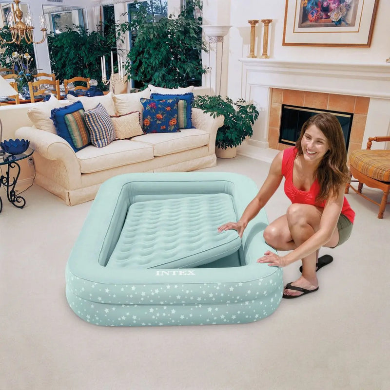 Laad afbeelding in galerijtoner, Een vrouw zet een Intex Kinder Kampeerluchtbed, een lichtblauw opblaasbaar kinderbed van Intex, neer in een lichte woonkamer met een witte bank en planten, om binnen kamperen extra leuk te maken voor kinderen.