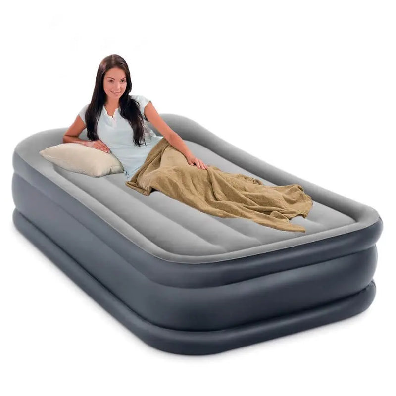 Laad afbeelding in galerijtoner, Een vrouw lacht naar de camera terwijl ze op het Intex Pillow Rest Deluxe Eenpersoons Luchtbed ligt, bedekt met een beige deken en kussen. Met de ingebouwde elektrische pomp is dit luchtbed snel en eenvoudig op te blazen.