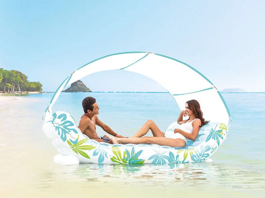 Een man en een vrouw ontspannen op de opblaasbare loungestoel Intex Tropical Canopy van Intex in ondiep, helder oceaanwater vlak bij het strand.