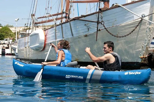 Twee personen peddelen in een Sevylor Adventure Kit Tweepersoons Opblaaskano met verstelbare stoelen bij een aangemeerde houten boot en glijden soepel over het kalme water.