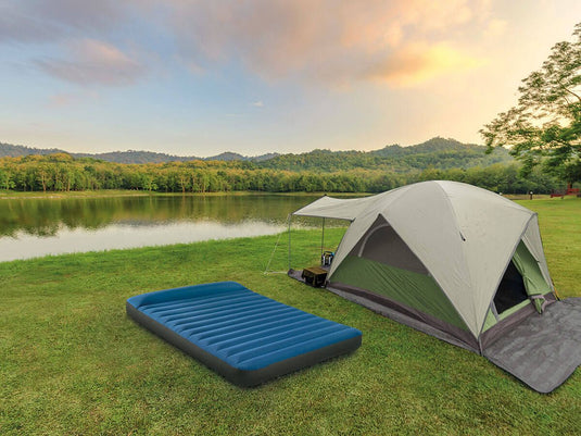 Een blauw Intex TruAire opblaasbaar twijfelaar kampeerluchtbed naast een opgezette tent beide staan op het gras aan een meer