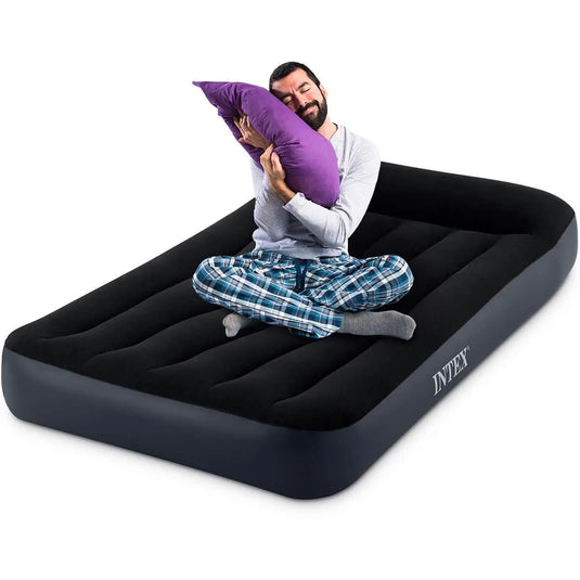 Een man in pyjama zit in kleermakerszit op een Intex Pillow Rest Classic Eenpersoons Luchtbed, knuffelt een paars kussen en kijkt tevreden.