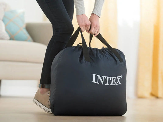Een persoon tilt een grote zwarte INTEK tas met handgrepen op in een woonkamer, ideaal voor het opbergen van uw Intex Comfort Plush Eenpersoons Luchtbed, dat is voorzien van geavanceerde Fiber-Tech® technologie.