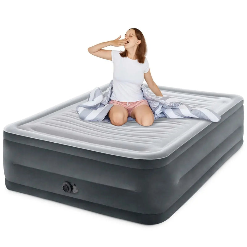 Laad afbeelding in galerijtoner, Een vrouw zit en gaapt op een grijs Intex Comfort Plush Tweepersoons Luchtbed, gedeeltelijk bedekt door een gestreepte deken. Het luchtbed is voorzien van Intex&#39;s Fiber-Tech® technologie.
